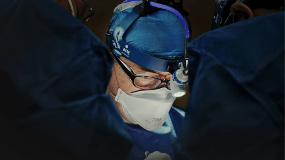 Photo of doctor with mask and surgical gown on lit by overhead light. THere are other people in the forefront but they are hidden in shadows